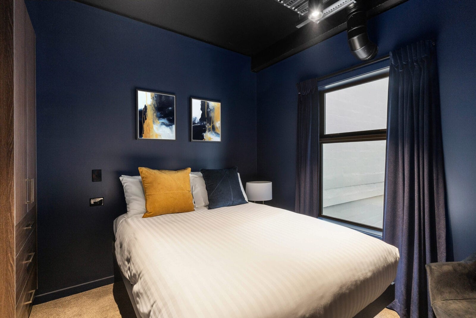 Cozy modern hotel room with dark navy walls, a double bed with striped white linens, and gold and navy throw pillows.