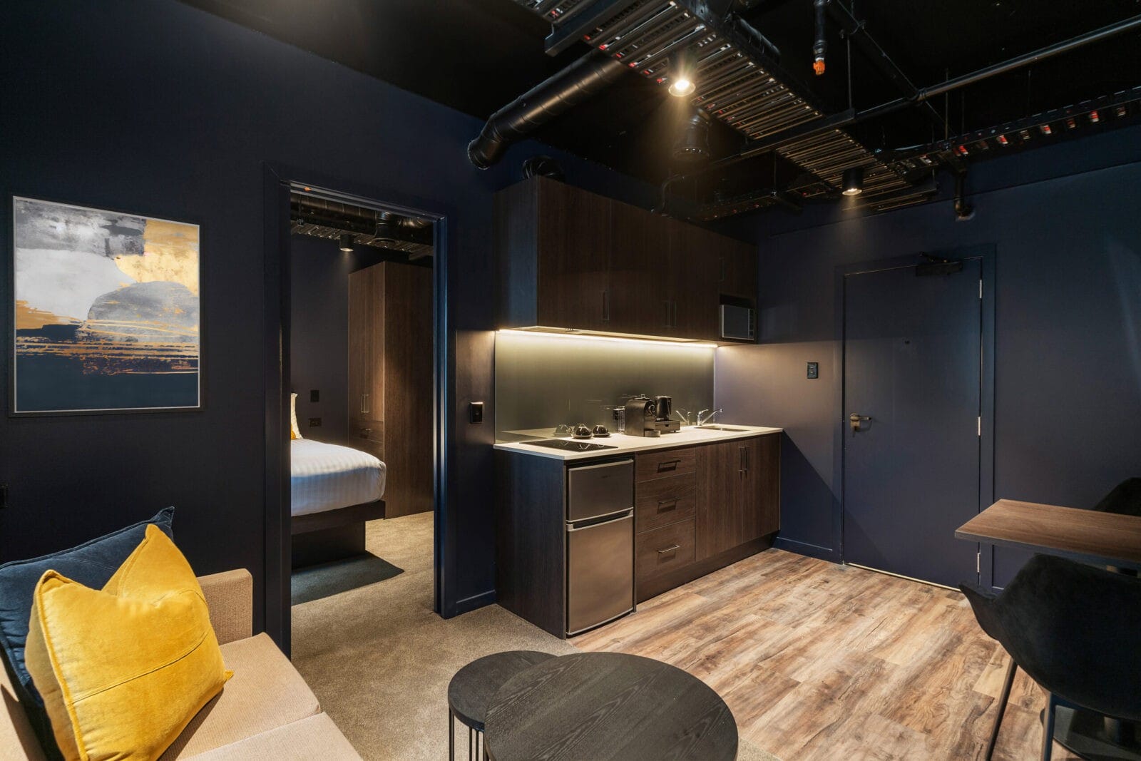 Modern hotel suite with a compact kitchenette, wood flooring, dark cabinetry, and a glimpse into a cozy bedroom with navy walls and soft lighting.
