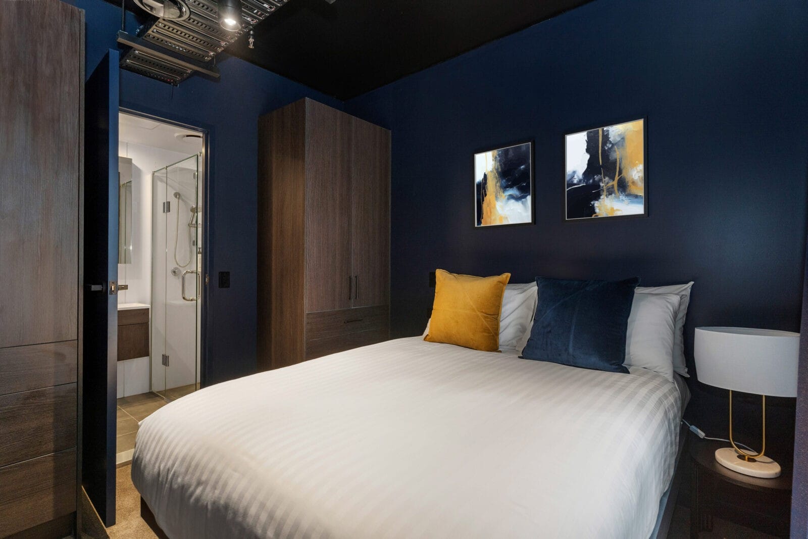 Contemporary hotel bedroom with dark blue walls, a queen bed with gold and navy cushions, and an open view of a sleek ensuite bathroom.