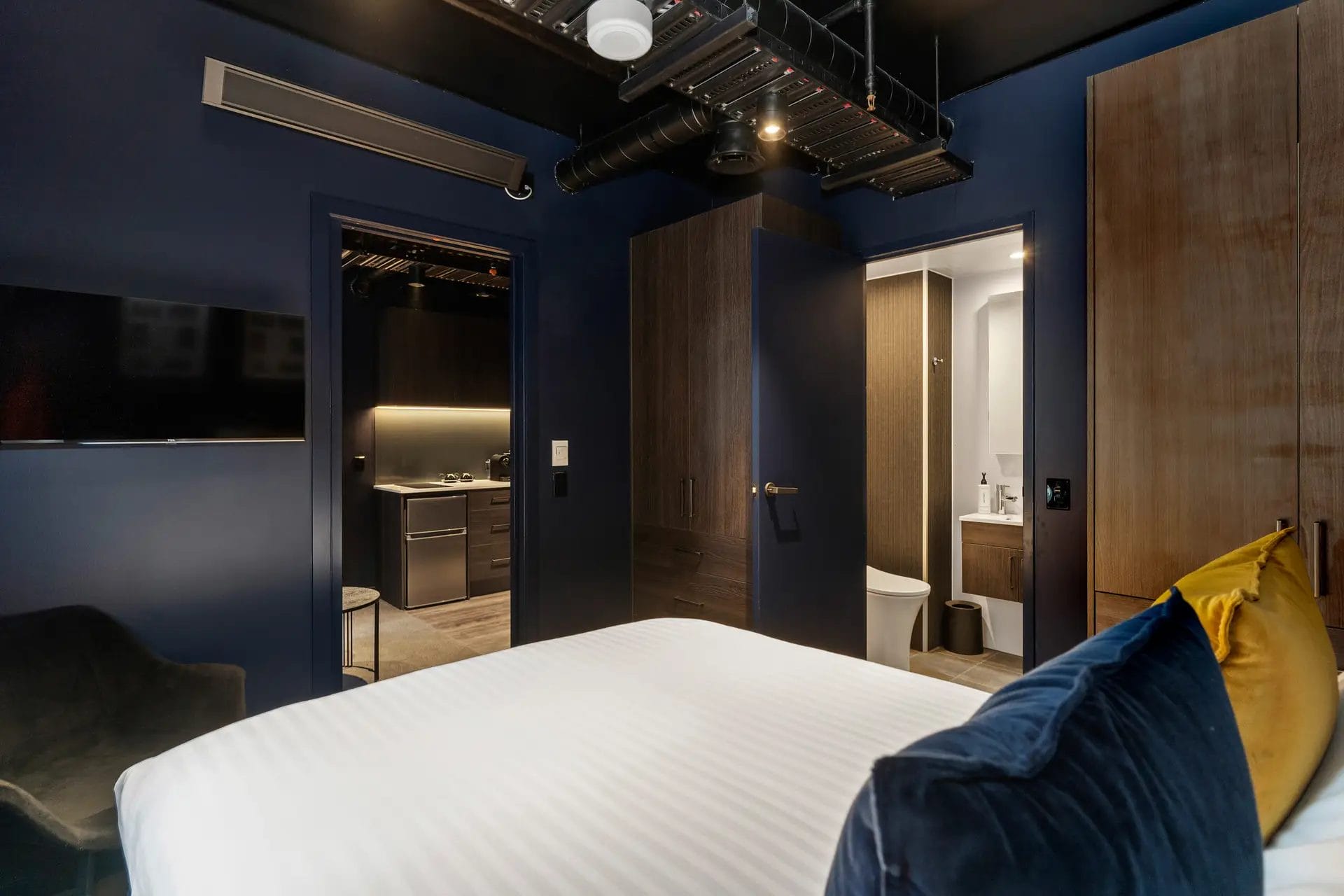 Perspective from hotel bed toward kitchenette and bathroom, featuring dark wood and navy tones.