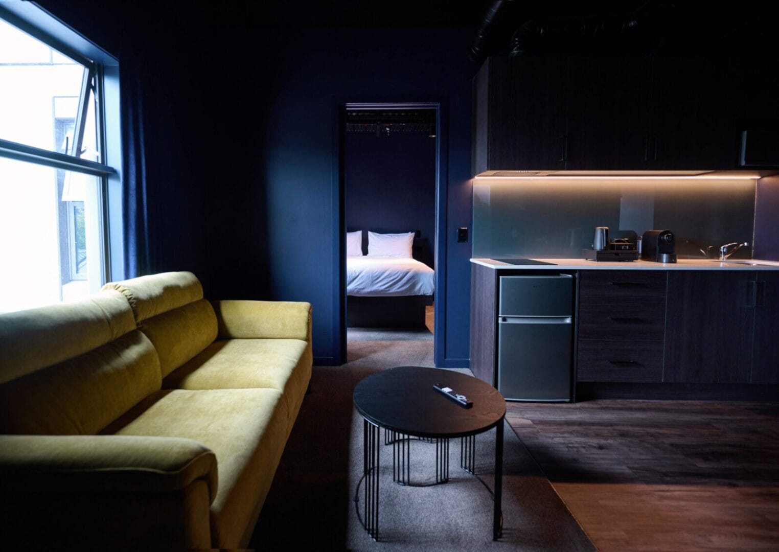 Modern hotel suite with yellow sofa, kitchenette, and view into a bedroom.