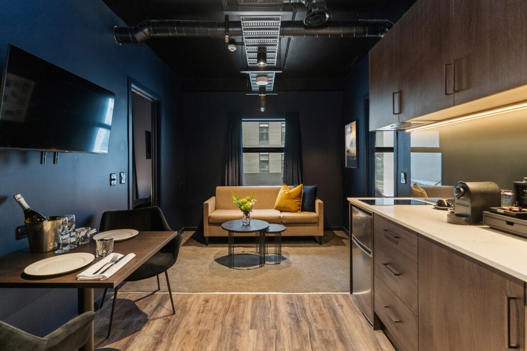 Contemporary hotel suite with kitchenette, dining setup, and beige sofa.