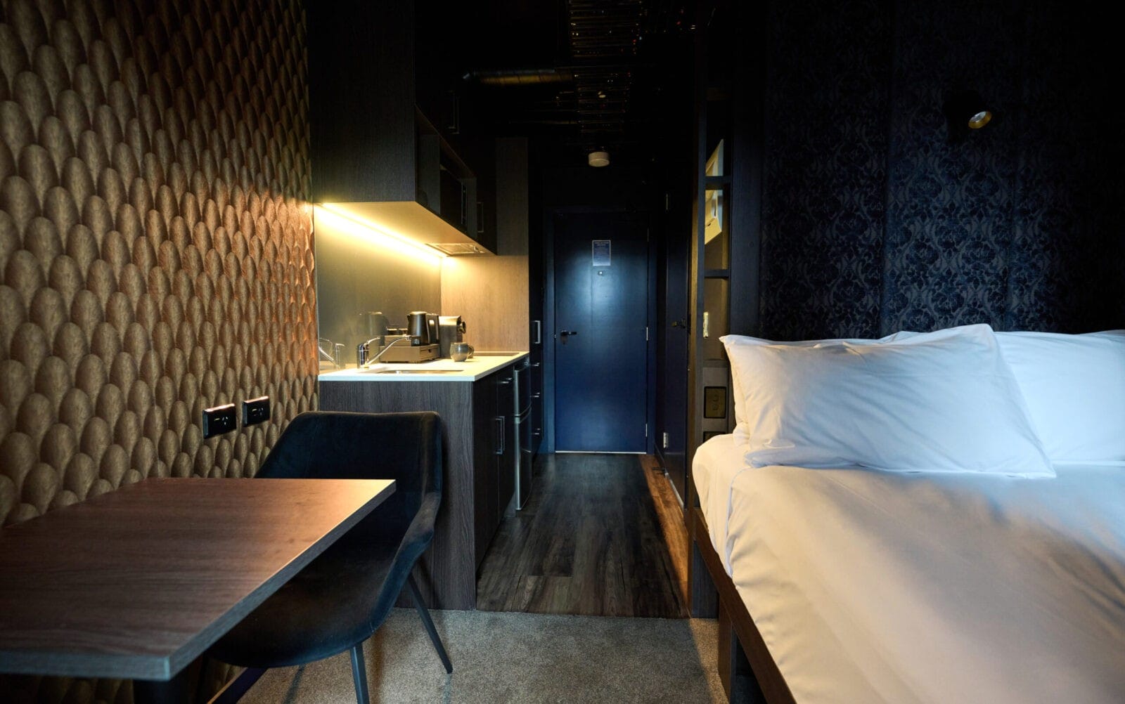 Modern hotel studio with a plush bed, compact kitchenette, and textured gold feature wall.