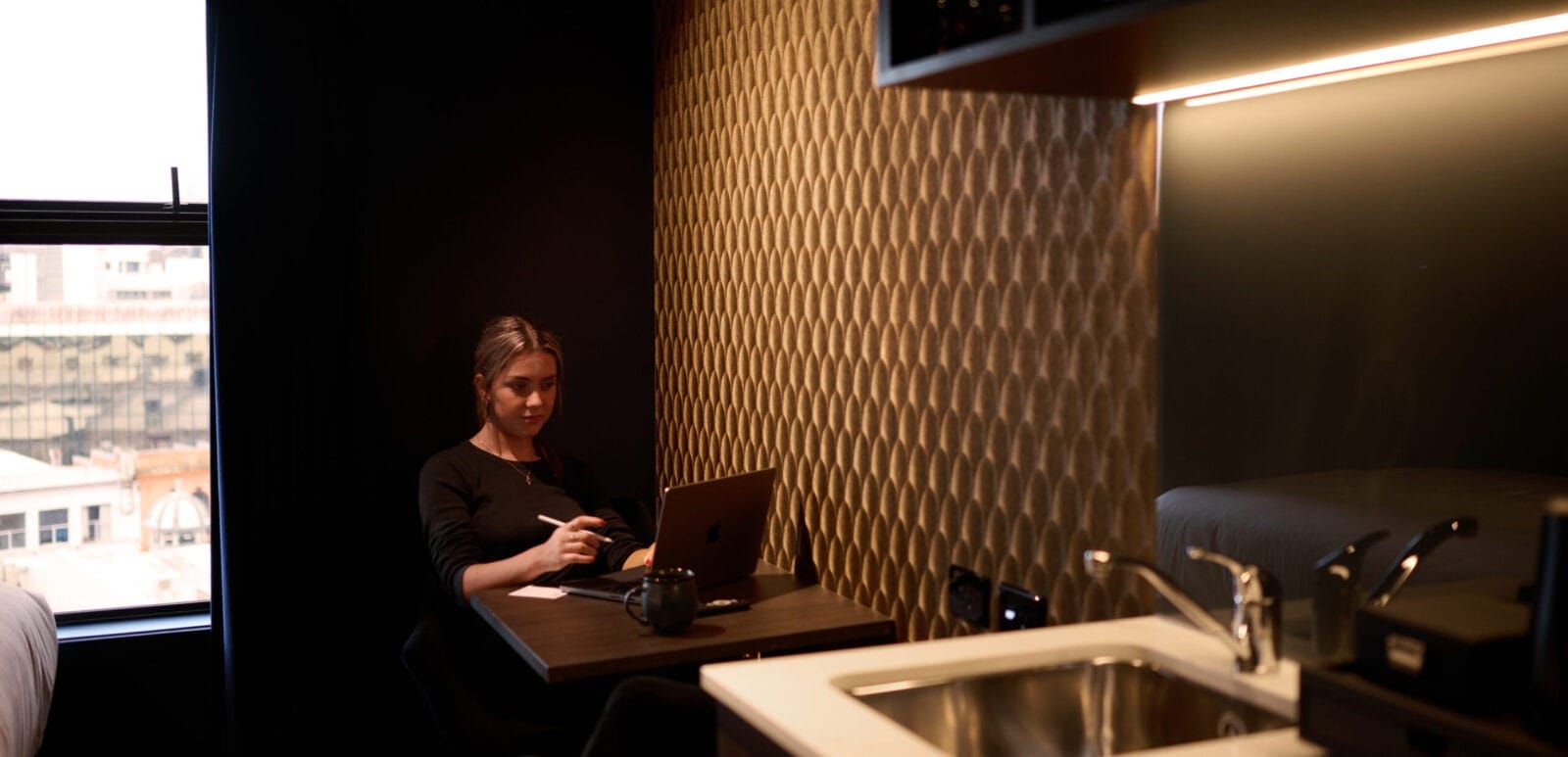 Woman working at a compact desk in a moody, modern hotel room with golden textured wallpaper and city views.