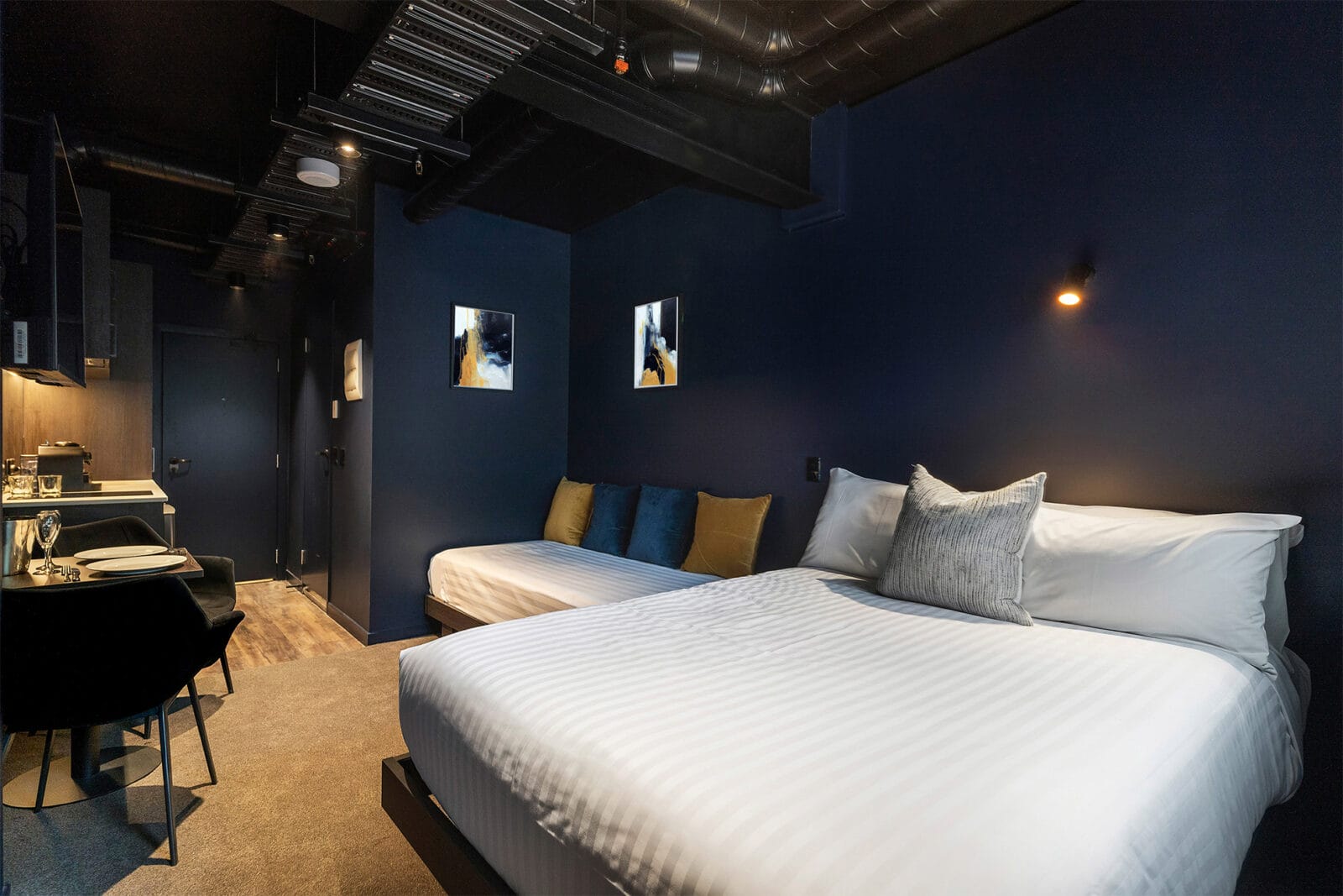 Stylish hotel room with dark navy walls, exposed ceiling, a queen bed, a single bed, and a compact dining area with a kitchenette.