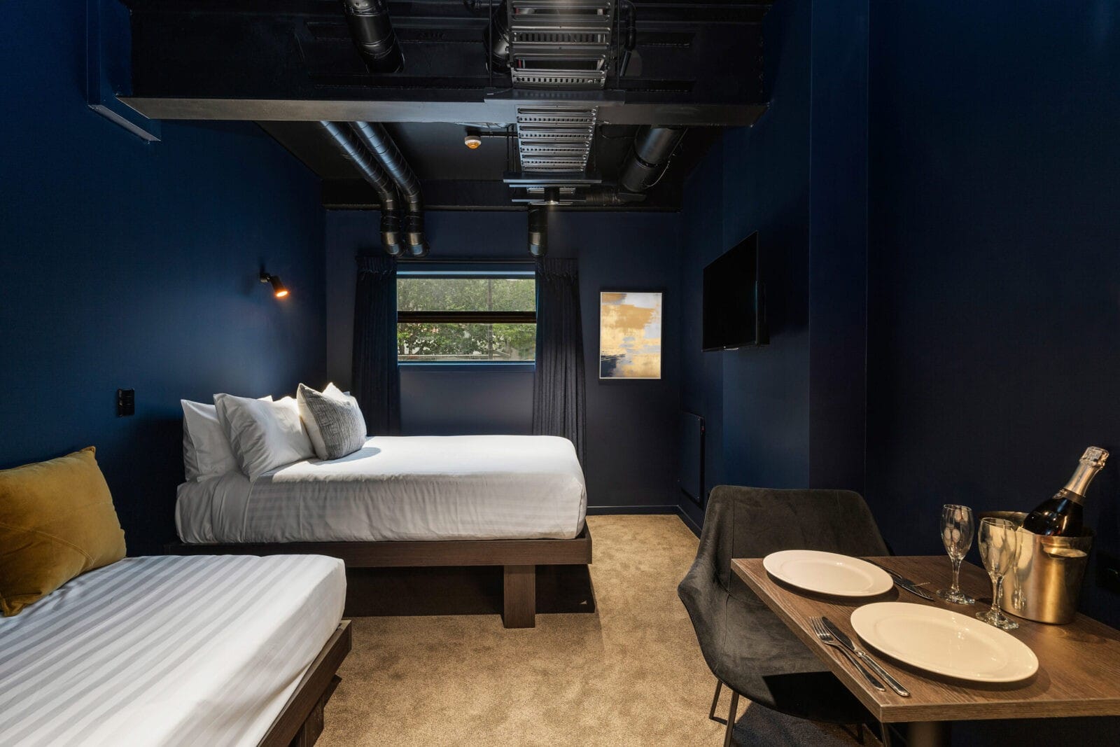 Modern hotel room with dark blue walls, a queen and single bed, window view, wall-mounted TV, and a dining table set with plates and champagne.