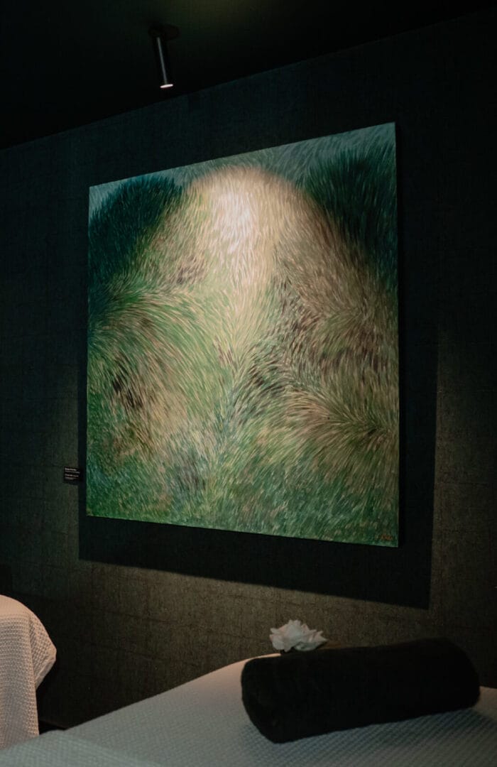 A softly lit spa room featuring a green abstract painting resembling wind-blown grass above a massage bed with a dark towel and white flower on top.