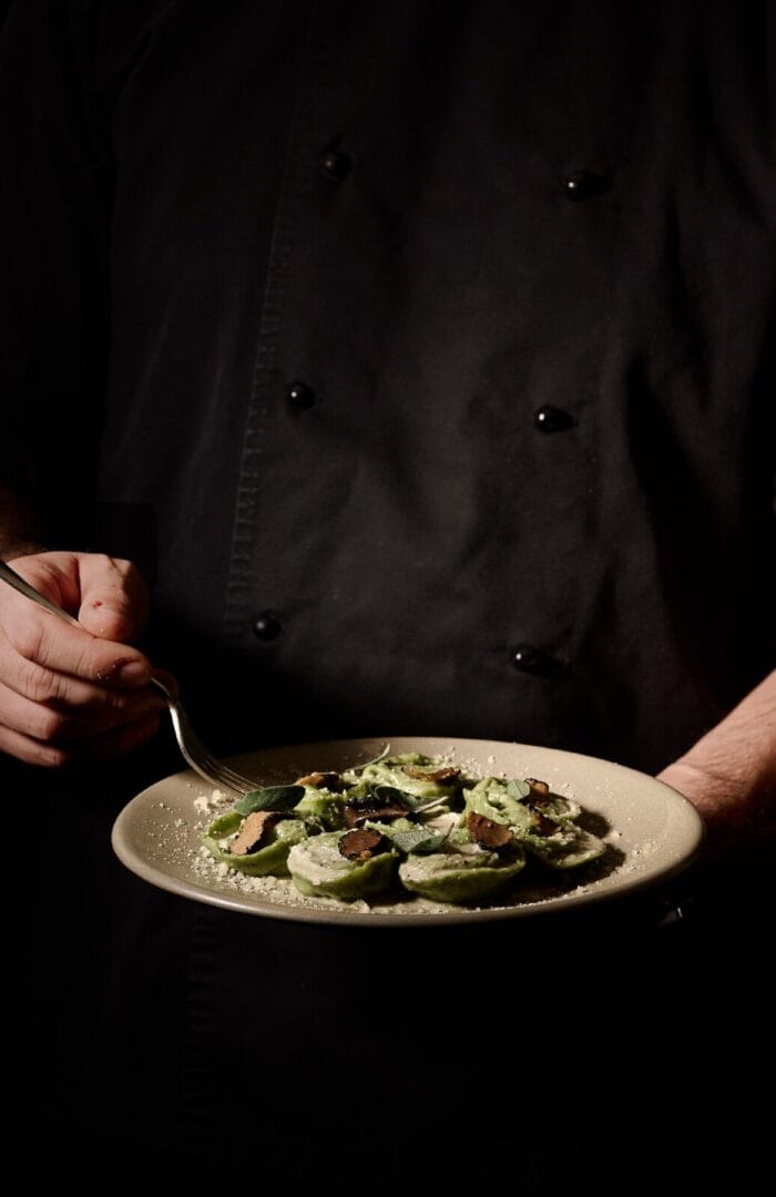 A chef in a black uniform holding a beige plate of handmade green ravioli, elegantly topped with shaved truffle, sage leaves, and grated cheese, under moody, warm lighting.
