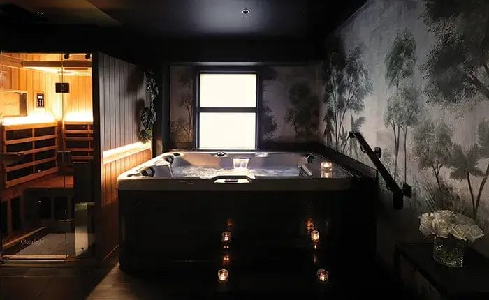 Luxurious private spa room with a warm-lit sauna, indoor hot tub, candlelight ambiance, and tranquil forest-themed wall mural.