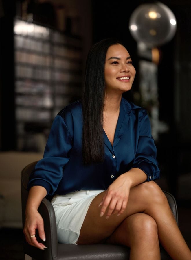 Smiling woman sitting in a modern lounge, wearing a satin blouse and white skirt.