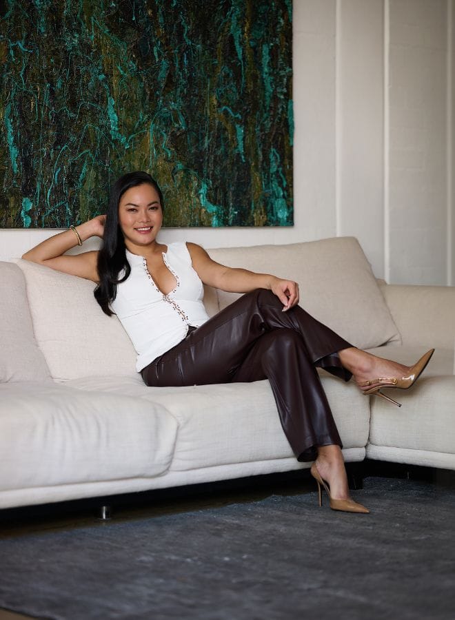 Woman sitting confidently on a modern beige sofa in a stylish interior, wearing a white top and leather pants.